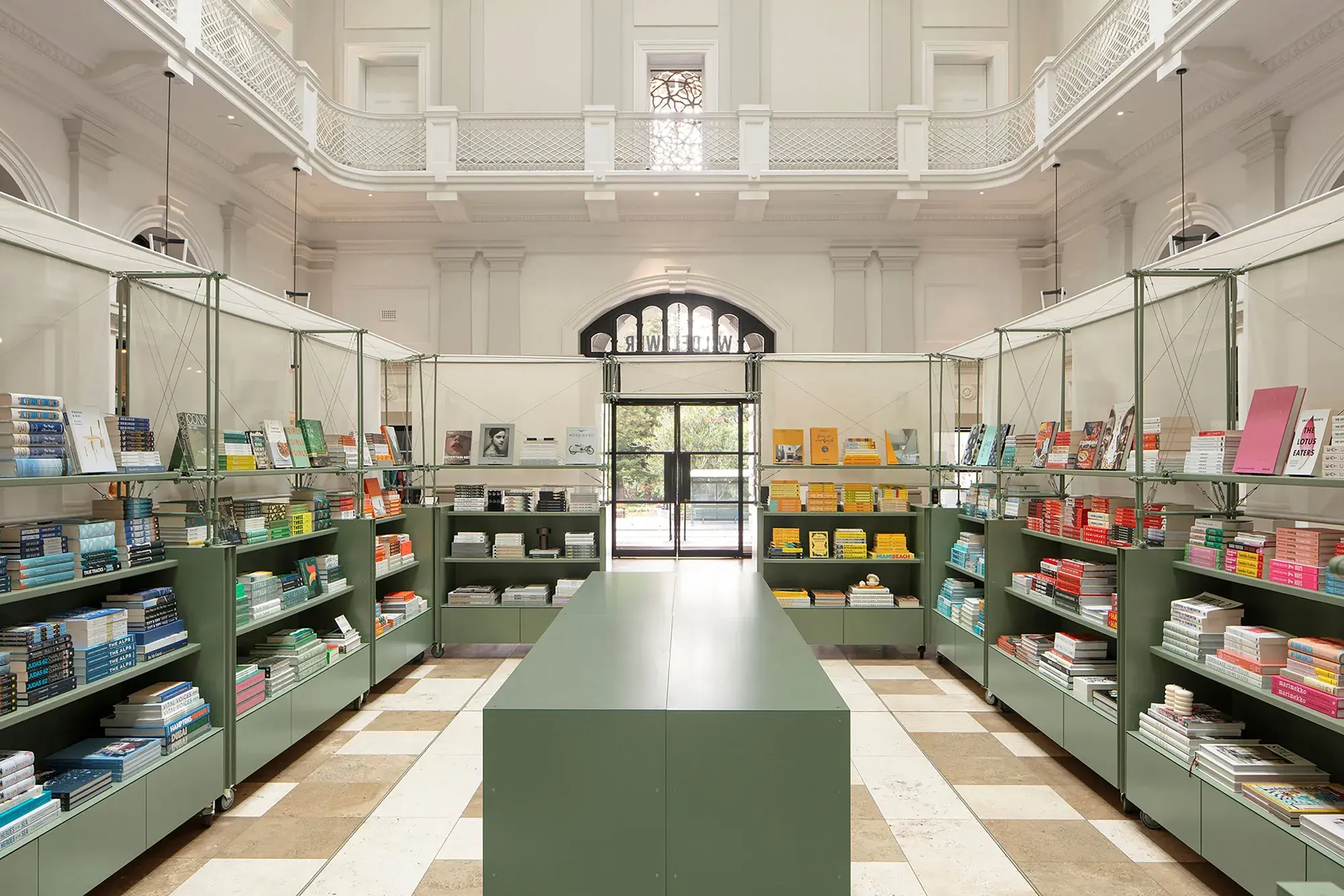A green island table sits in the middle of a room surrounded by bookshelves which are also green. There is a second story visible with a balcony to look down on this area.
