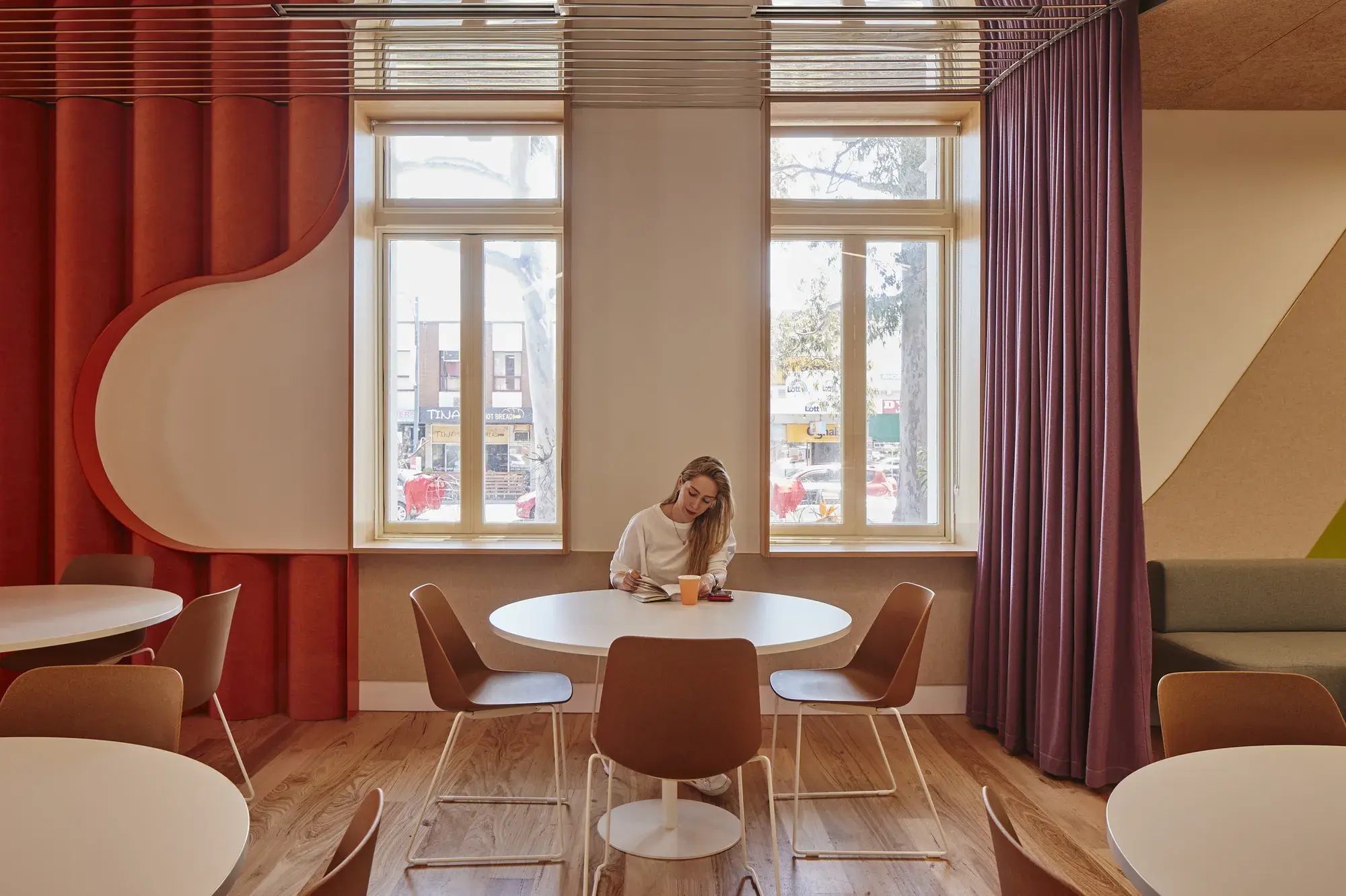 A woman sitting at a table in front of two windows. The features of the room are a red/orange colour and there are cream highlights on the walls.