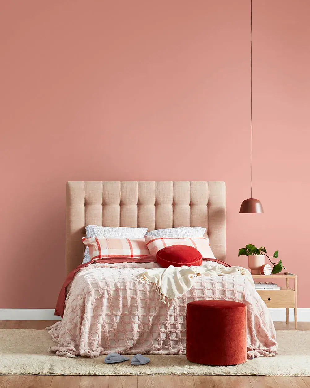 A bedroom featuring a bed with a fabric headboard and blankets that are pink toned. There is a pink hanging lamp by the bed and the wall is painted pink. There is a dark pink/red ottoman and a bedside table with a potted plant on it by the bed.
