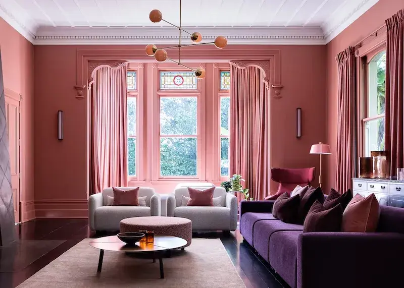 A sitting room with purple couches and light coloured sitting chairs. The walls and trims are a dusty pink colour and feature a feature arch around the large windows. 