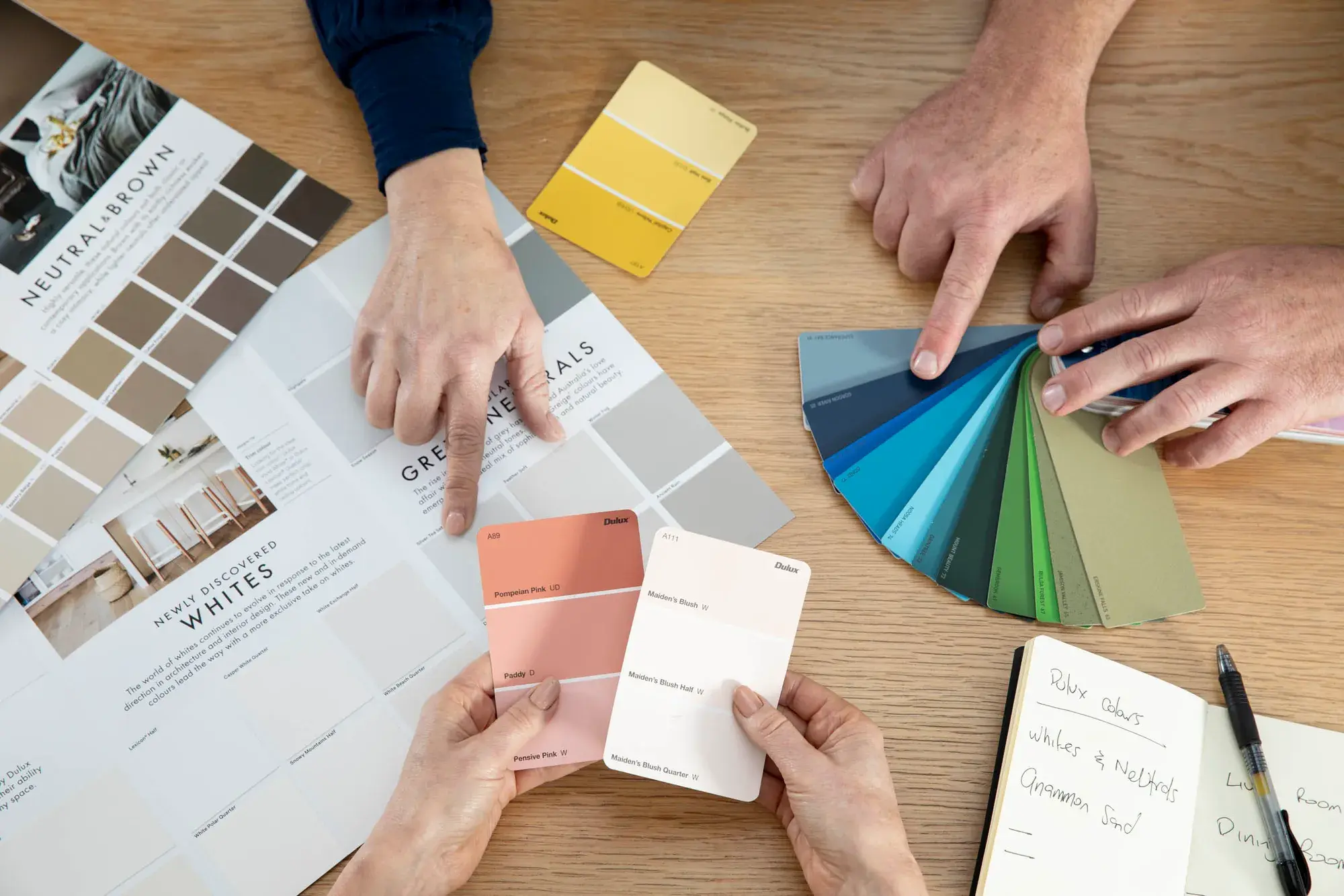Three hands holing Dulux colour chip cards displaying orange, whites and yellow schemes. 