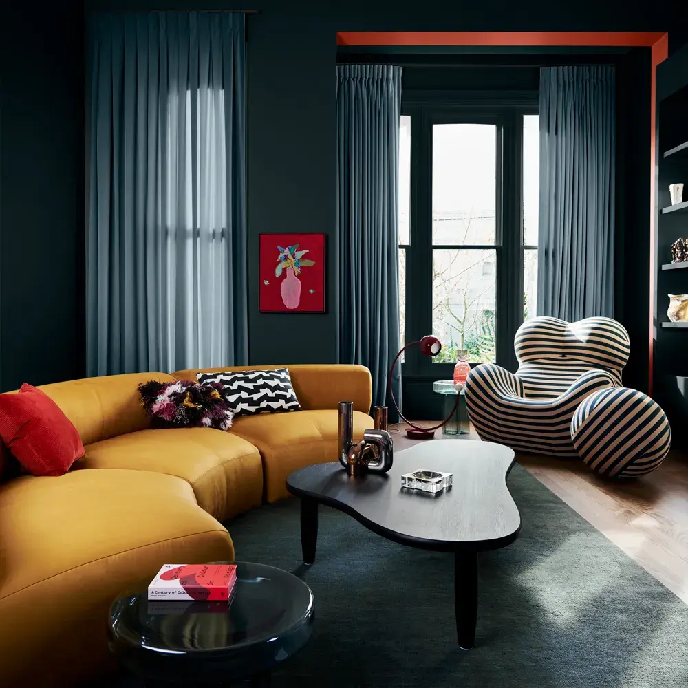 Bright yellow couch in dark room decorated with quirky striped chair and various decorative items.