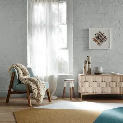 Living area with chair and blanket. Buffet with decorative items and window with sheer curtains.