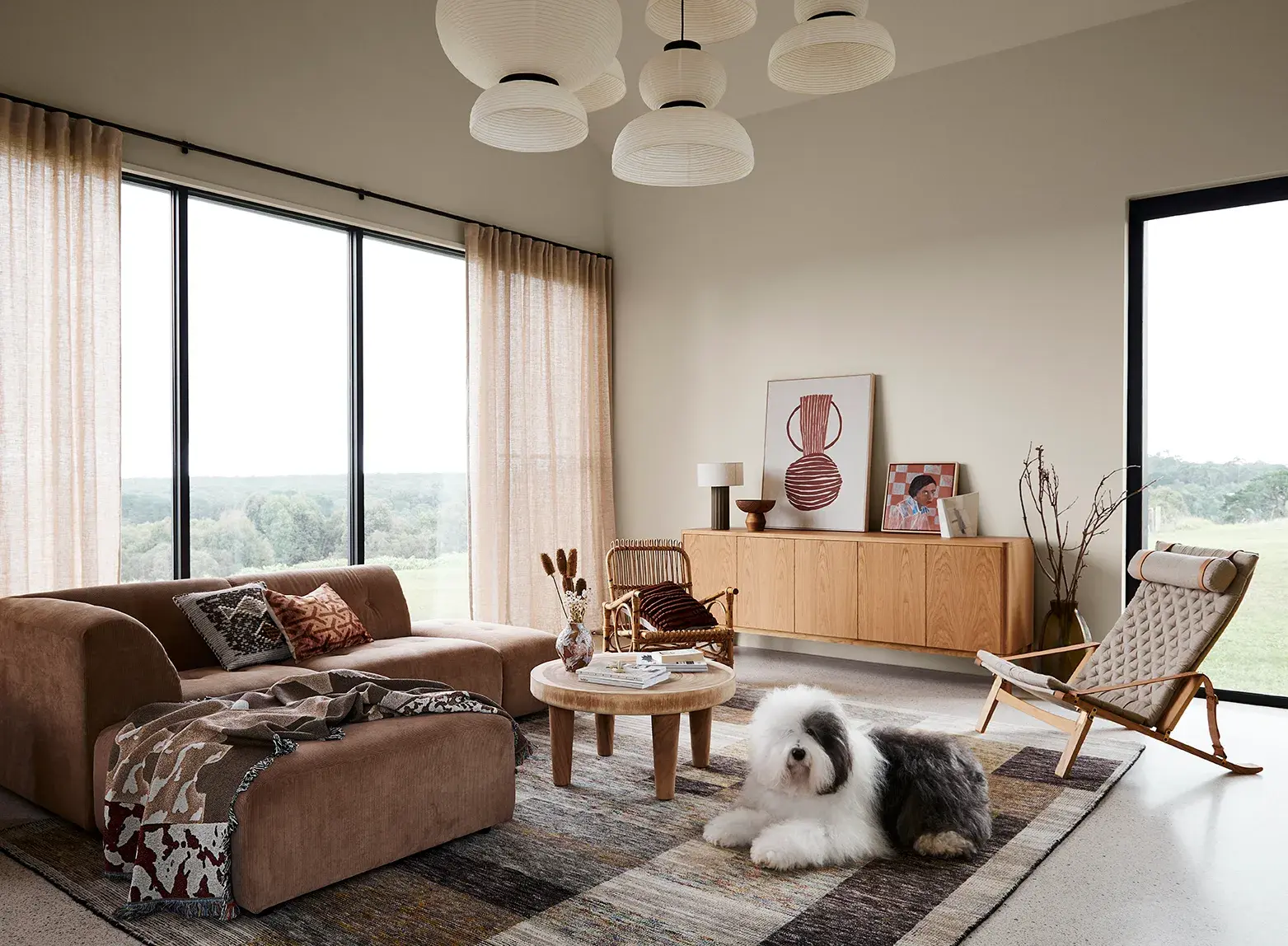 The Dulux Sheepdog lies down in a neutral-coloured living room on a rug in front of an L-shaped brown couch.