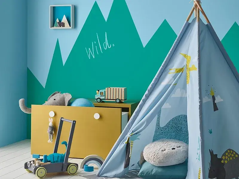 Dulux upcycling & decor project - A children's playroom with blue tent that has a children's squishy chair inside it. Next to it are yellow draws that are open with some toys spilling out. The wall is painted with a green and blue mountain pattern.