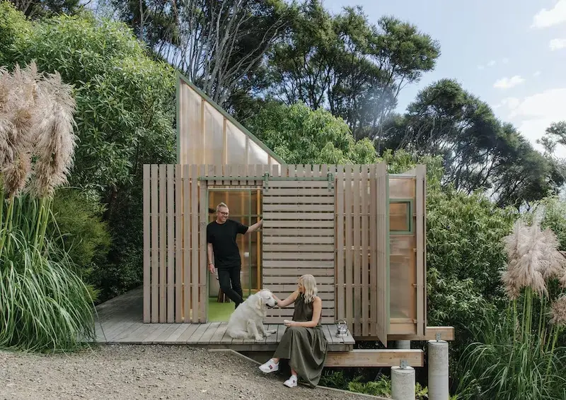 A woman is sitting on a wooden deck of a small structure with a golden retriever dog. There is a man standing in the doorway behind her smiling at them. 