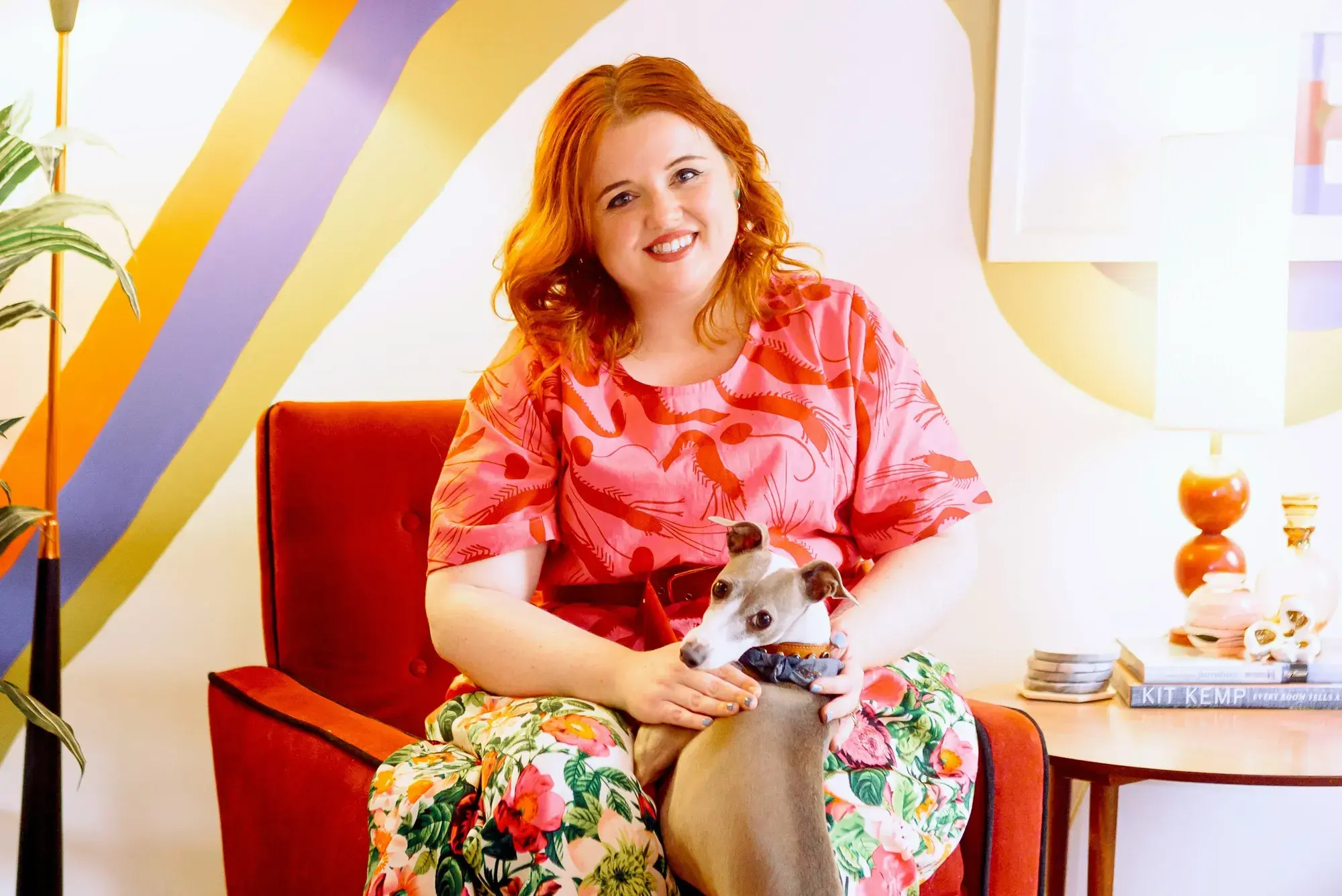Evie kemp sits on a chair in front of a rainbow wall with her dog jumping up on her lap.