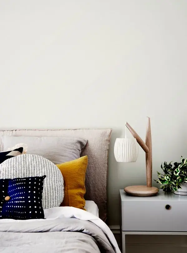 Close up of bedhead with multiple coloured cushions and a bedside table with a wooden lamp and plant 