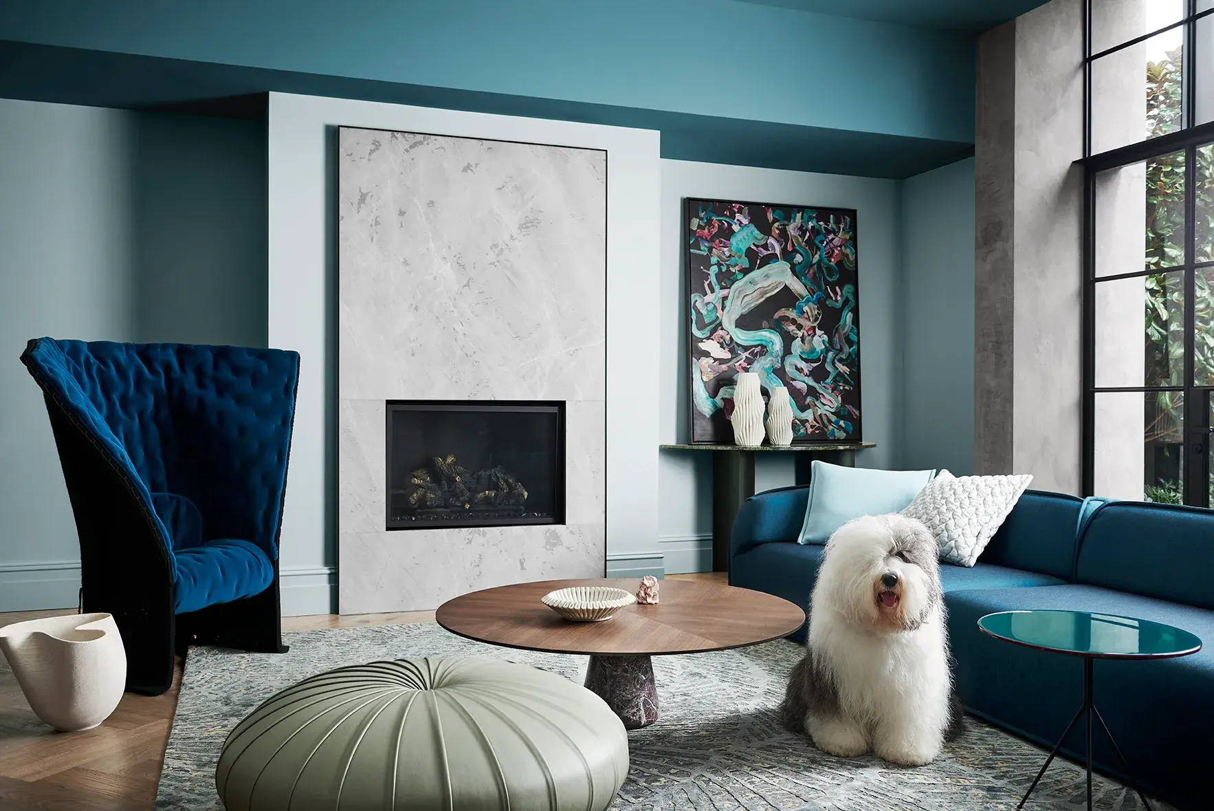 The Dulux sheepdog sits in a living room with blue-toned walls and a marble fireplace.