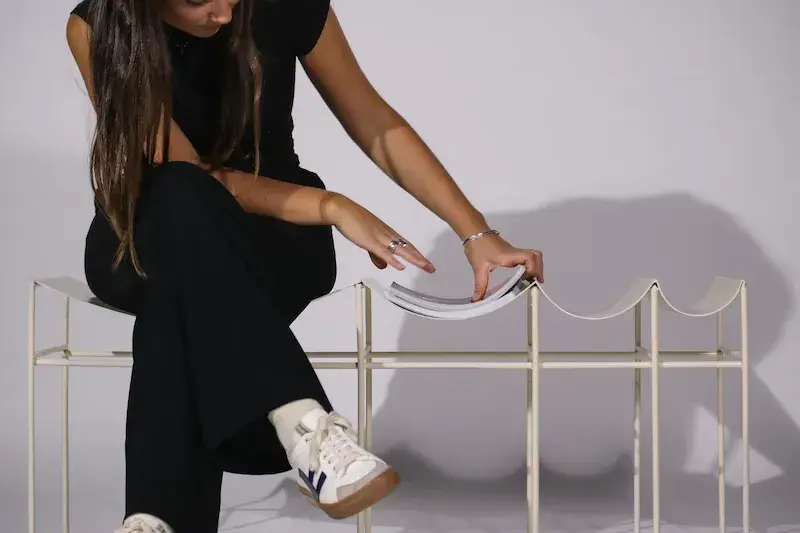 A woman sitting on the canopy bench which is a cream coloured bench with curved seat spaces that slowly get smaller as you move along the bench
