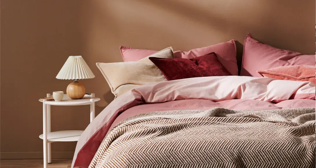 Interior bedroom zoomed in on bed featuring pink bed spread and red pillows against a beige feature wall.